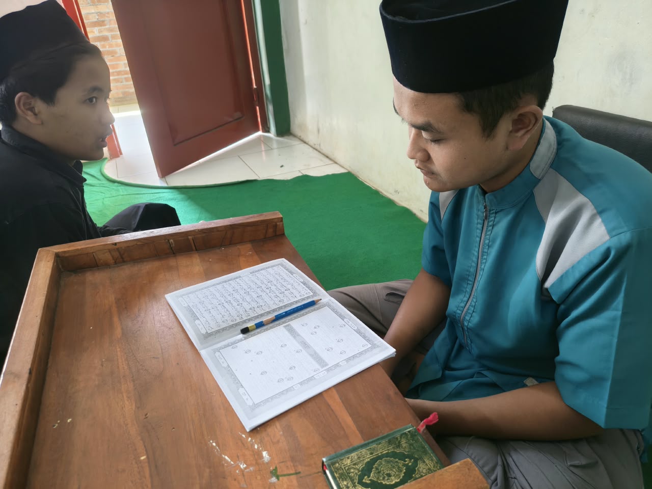 Santri menyetorkan hafalan, musyrif menyimak dengan buku hafal tulis terbuka